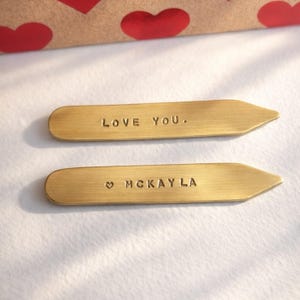 Two brass collar stays, one engraved "LOVE YOU." and the other "♡ MCKAYLA", rest on a white surface with a red heart-patterned box in the background.
