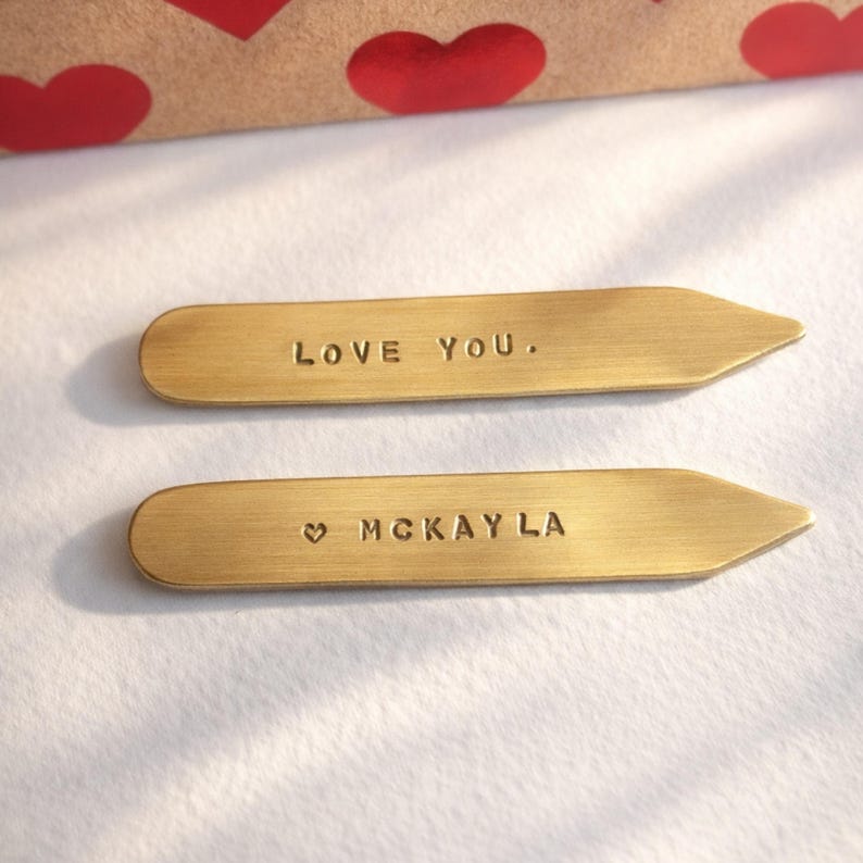 Two brass collar stays, one engraved "LOVE YOU." and the other "♡ MCKAYLA", rest on a white surface with a red heart-patterned box in the background.