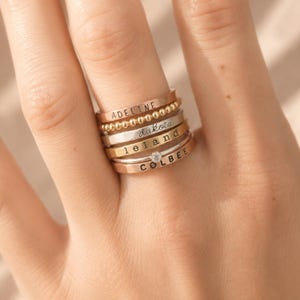 Puede incluir: Un conjunto de seis anillos de metal en tonos oro rosa y plata. Los anillos presentan varias texturas, incluyendo una banda de cuentas, una banda con una pequeña piedra clara y bandas planas grabadas con los nombres ADELINE, dakota, leland y COLBEE.