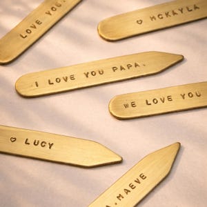 Six brass collar stays are scattered on a light surface, each engraved with different text like "LOVE YOU PAPA" and "WE LOVE YOU."