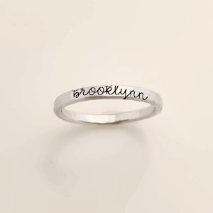 May include: A silver ring with the name "Brooklyn" engraved in a cursive font. The ring is a simple band design, likely made of sterling silver or a similar metal. The ring is shown against a neutral background.