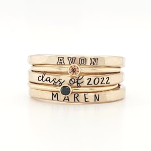 A five piece yellow gold class ring set. The set consists of three hand made, lightly hammered rings with the word 'Avon' engraved on one, '2022' engraved on one and the name 'Maren' engraved on one. All are engraved in black. The rings are separated by yellow gold stacking birthstone rings, one yellow and one black. The rings are on featured against a white background.