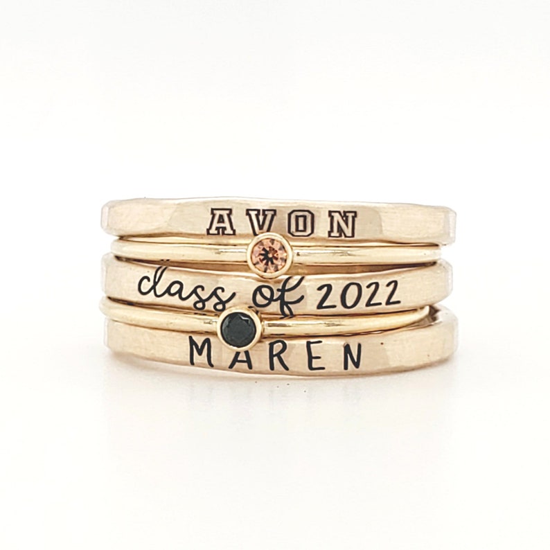 A five piece yellow gold class ring set. The set consists of three hand made, lightly hammered rings with the word 'Avon' engraved on one, '2022' engraved on one and the name 'Maren' engraved on one. All are engraved in black. The rings are separated by yellow gold stacking birthstone rings, one yellow and one black. The rings are on featured against a white background.