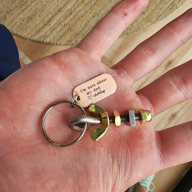May include: A metal keychain featuring a collection of nuts and bolts, with a small copper tag that reads "I'm nuts about my dad. Charley". The keychain includes a silver key ring and a gold-colored wing nut.