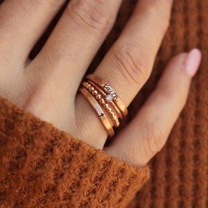 A rose gold ring set consisting of a handmade ring engraved with the word "faith" in black, a handmade ring engraved with a cross in black, a rose gold beaded ring and a thin clear August birthstone. The set is worn by a woman in a brown sweater.