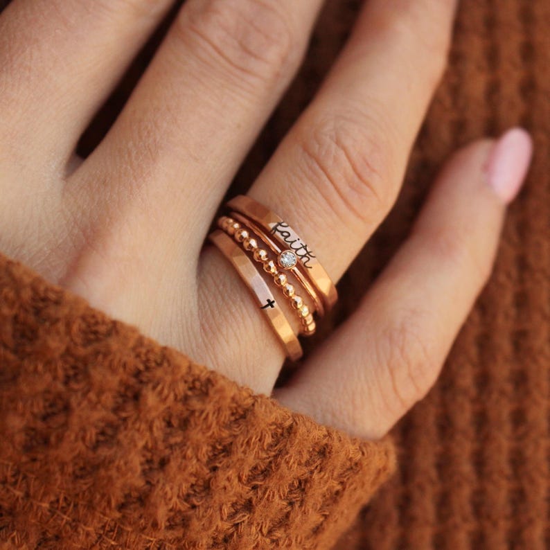 A rose gold ring set consisting of a handmade ring engraved with the word "faith" in black, a handmade ring engraved with a cross in black, a rose gold beaded ring and a thin clear August birthstone. The set is worn by a woman in a brown sweater.