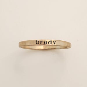 May include: A thin, gold-toned metal ring with a hammered texture. The word "brady" is stamped in black, lowercase, sans-serif lettering across the front center of the band. The ring has a simple, narrow profile and a metallic finish.