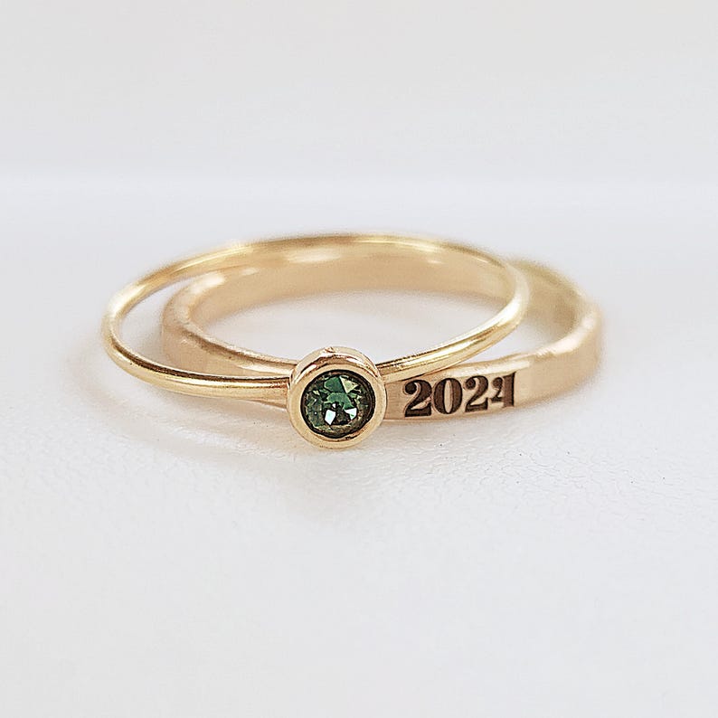 One gold ring with a green gem and one gold ring that says 2024