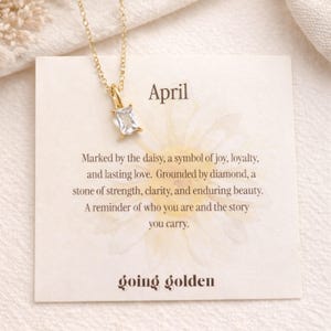 May include: A gold-colored necklace with a rectangular, clear gemstone pendant. The necklace hangs over a card that reads "April" and includes text about the daisy and diamond. The card also has the words "going golden".