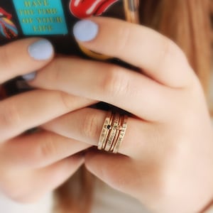 A five piece yellow gold class ring set. The set consists of three hand made, lightly hammered rings with the word 'Avon' engraved on one, '2022' engraved on one and the name 'Maren' engraved on one. All are engraved in black. The rings are separated by yellow gold stacking birthstone rings, one yellow and one black. The rings are on the hand of a woman wearing a white shirt, holding a phone with bright colored stickers on it.