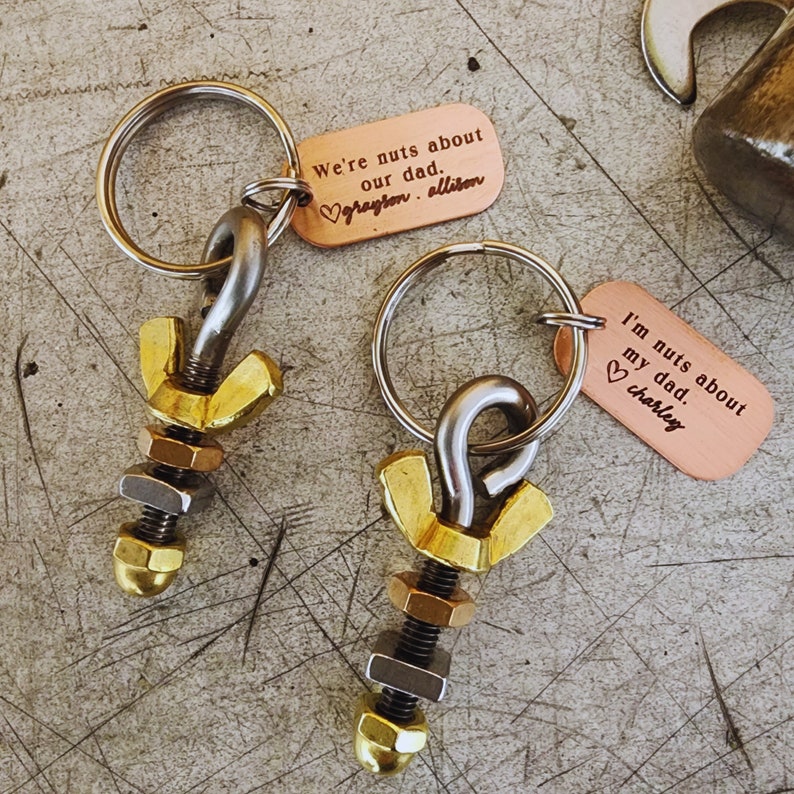 May include: Two keychains with metal hardware and copper tags. The tags read "We're nuts about our dad" and "I'm nuts about my dad," with names below. The keychains feature a mix of gold, silver, and bronze-colored metal components.
