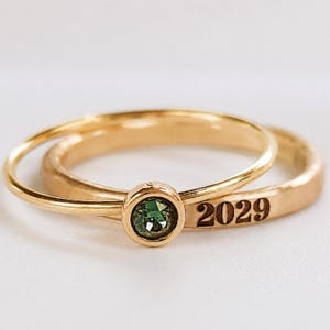 May include: A gold ring set consisting of two bands. One band features a round, bezel-set green gemstone. The second band is engraved with the number 2029. Both rings have a polished finish and are displayed together in a stacked arrangement.