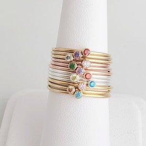 Puede incluir: Pila de anillos de oro, plata y oro rosa con peque&ntilde;as piedras preciosas de colores. Cada anillo tiene una piedra preciosa de color diferente, incluyendo rojo, verde, azul, morado y transparente.