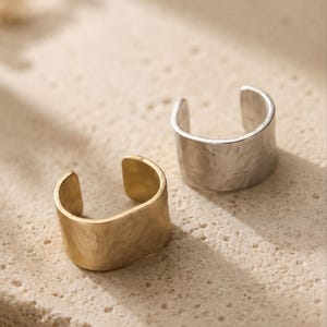 May include: Two open-ended ear cuffs, one in a gold-tone finish and one in a silver-tone finish. Both cuffs feature a wide, band-style design with a textured, hammered surface and a smooth, polished interior edge, creating a simple, open circular shape.