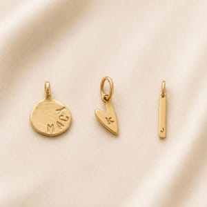 Op de afbeelding: Drie goudkleurige metalen bedels, bestaande uit een ronde hanger met de inscriptie MACY, een hartvormige hanger met de letter K en een slanke, rechthoekige staafhanger met de letter J, elk voorzien van een klein verbindingsringetje.