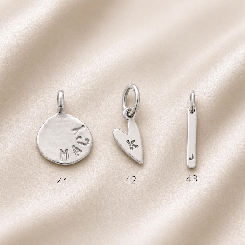 Three silver charms are shown: a round disc with "MACY" stamped on it, a heart with "K" stamped on it, and a plain rectangular bar.