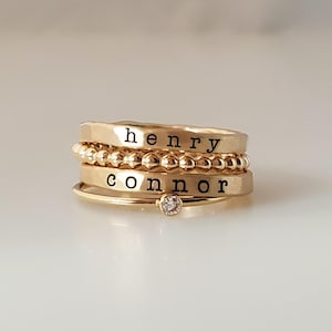 A stack of four gold-colored rings with engraved names henry and connor, a beaded band, and a crystal solitaire band.