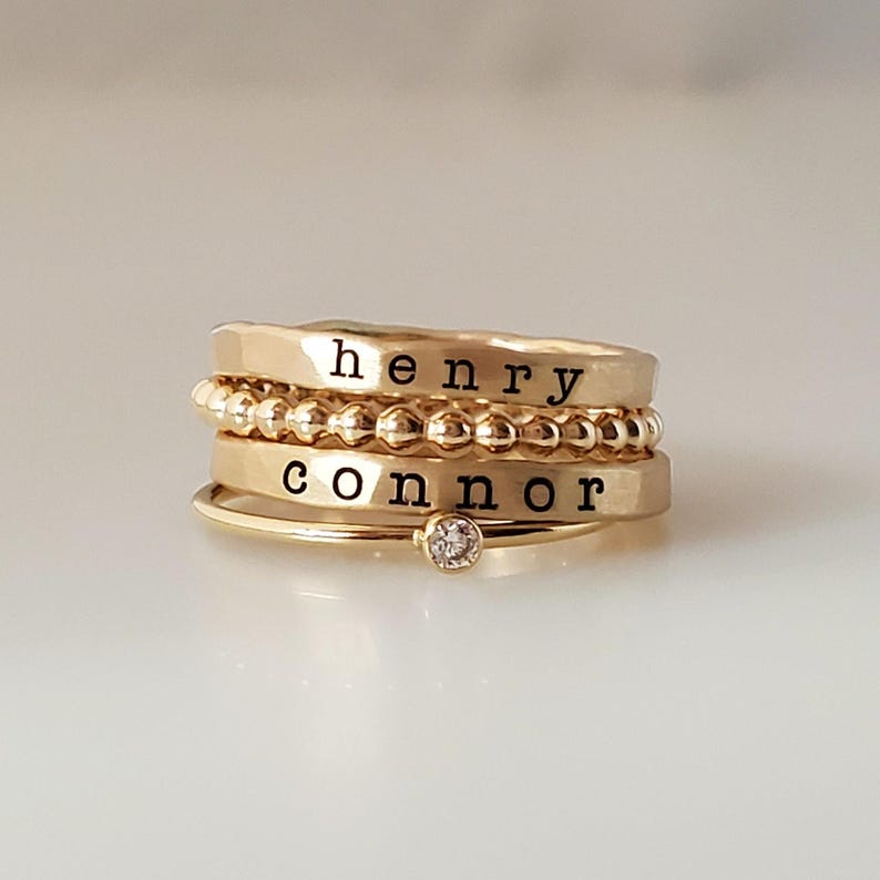 A stack of four gold-colored rings with engraved names henry and connor, a beaded band, and a crystal solitaire band.