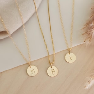 May include: Three gold-coloured necklaces with circular pendants, each bearing a single initial: "M", "B", and "K". The pendants hang from delicate chains, resting on a light-coloured surface. The necklaces are simple and elegant.
