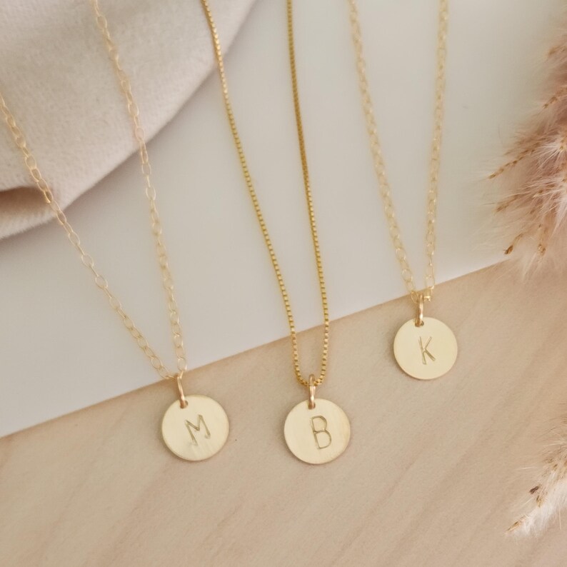 May include: Three gold-coloured necklaces with circular pendants, each bearing a single initial: "M", "B", and "K". The pendants hang from delicate chains, resting on a light-coloured surface. The necklaces are simple and elegant.