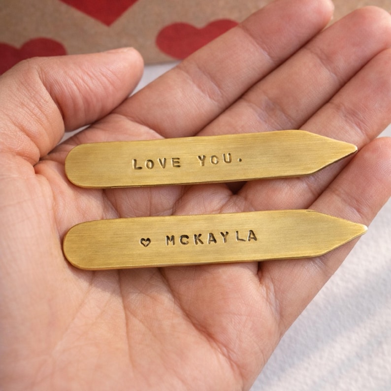 Two brass collar stays, one engraved "LOVE YOU." and the other "♡ MCKAYLA", are held in open hands, with a red heart-patterned box in the background.