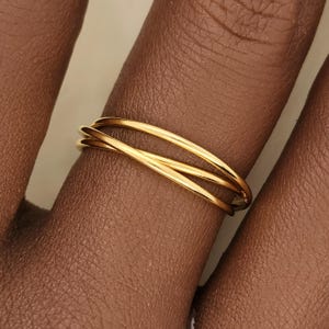 May include: A gold-colored ring with a triple-band design. The ring is worn on a finger, showcasing its intertwined bands. The ring's polished surface reflects light, highlighting its smooth, curved lines. The jewelry piece is a simple yet elegant accessory.