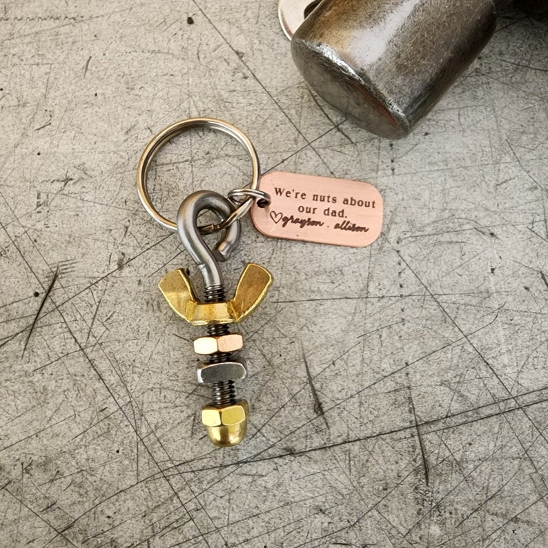 May include: A handmade keychain featuring a metal bolt design with a brass wing nut and a copper tag. The tag is engraved with the words "We're nuts about our dad." and a heart symbol, followed by names. The keychain is attached to a silver keyring.