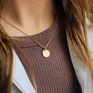 May include: A gold necklace with a circular pendant. The pendant has the letter "B" in the centre. The necklace is worn against a brown ribbed top and a white shirt.
