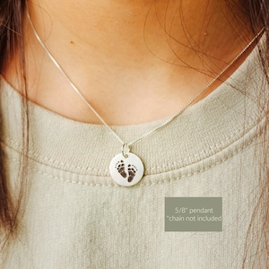 May include: Silver pendant necklace featuring a small, round charm with an engraved image of baby footprints. The pendant is 5/8 inch in diameter and hangs from a delicate silver chain. The necklace is displayed on a person wearing a light green shirt.