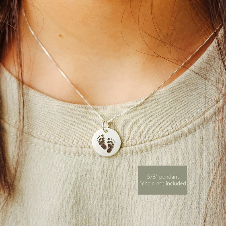May include: Silver pendant necklace featuring a small, round charm with an engraved image of baby footprints. The pendant is 5/8 inch in diameter and hangs from a delicate silver chain. The necklace is displayed on a person wearing a light green shirt.