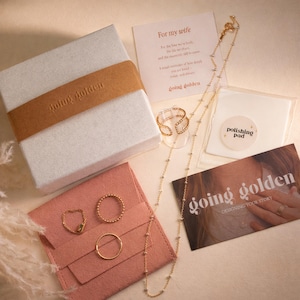 A white gift box with a brown ribbon, a white card with red text, a polishing pad, and various gold-colored jewelry items on a light brown surface.