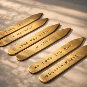 Six brass collar stays are arranged in two rows on a light surface, each engraved with different personalized messages.