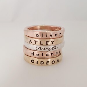 May include: A stack of five personalized rings with the names Oliver, Atley, Sawyer, Delaney, and Gideon engraved on them. The rings are made of different metals, including gold, silver, and rose gold.