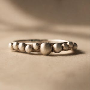 Puede incluir: Un anillo de tono plateado que presenta una serie de pequeñas esferas redondeadas de distintos tamaños dispuestas a lo largo de la banda exterior. El metal tiene un acabado mate y texturizado, con esferas colocadas una junto a la otra.