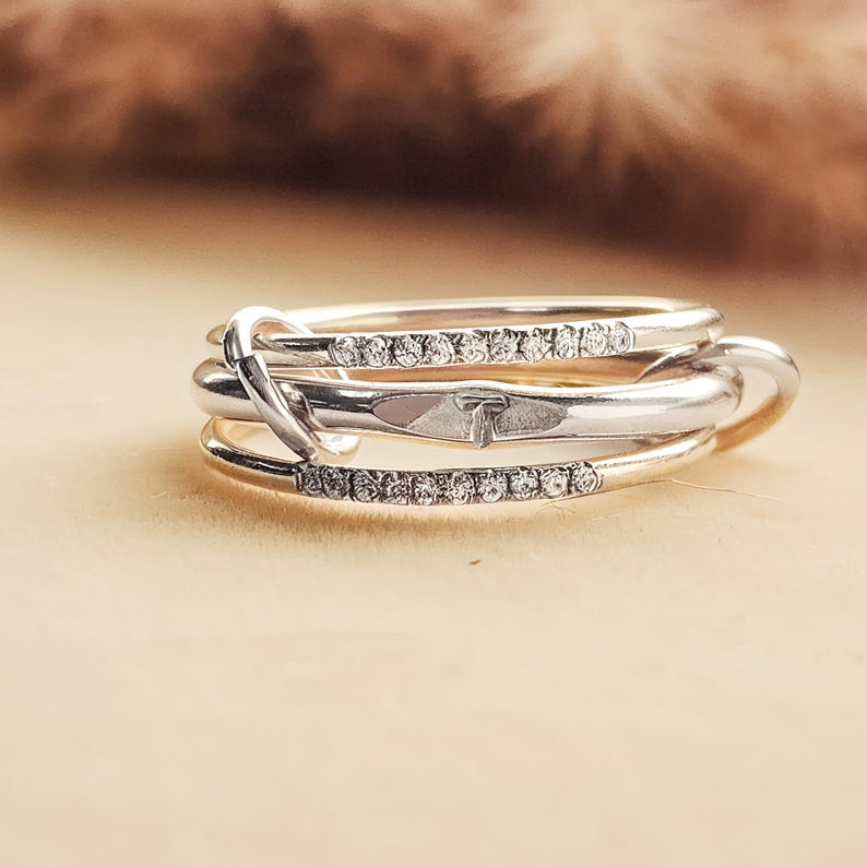 Pode incluir: Um anel de prata com tr&ecirc;s faixas. Duas faixas s&atilde;o adornadas com pequenas pedras preciosas transparentes. As faixas s&atilde;o entrela&ccedil;adas com um design de n&oacute;. O anel &eacute; exibido em uma superf&iacute;cie bege clara com um fundo marrom desfocado.
