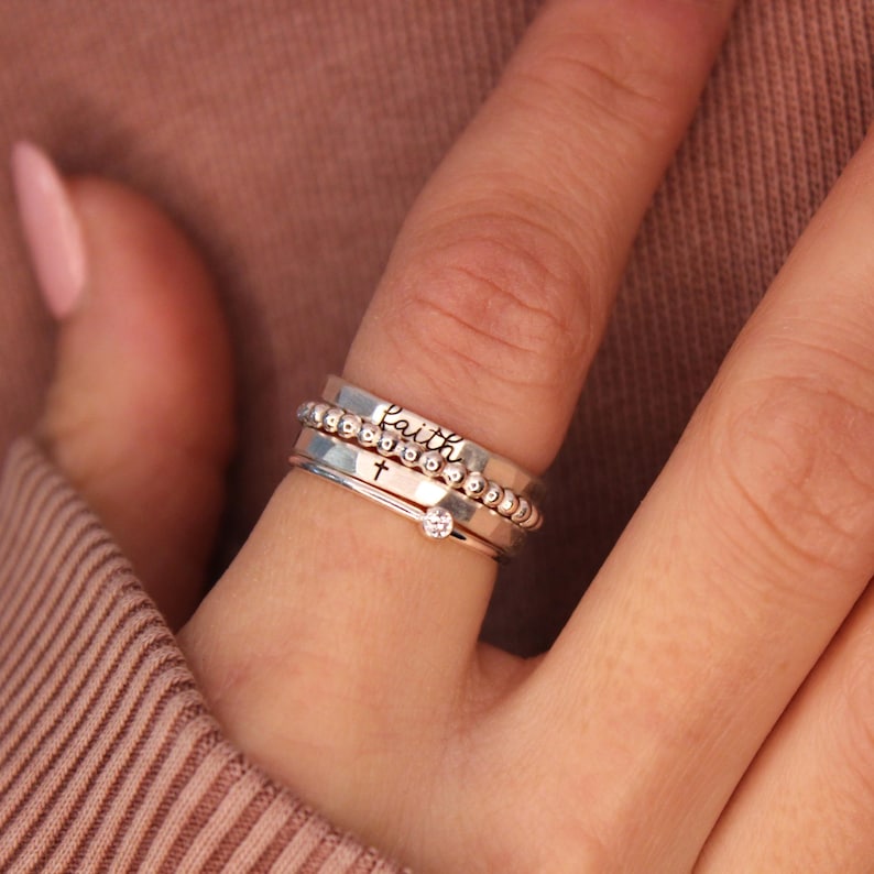 A silver ring set consisting of a handmade ring engraved with the word "faith" in black, a handmade ring engraved with a cross in black, a silver beaded ring and a thin clear August birthstone. The set is worn by a woman in a pink sweater.