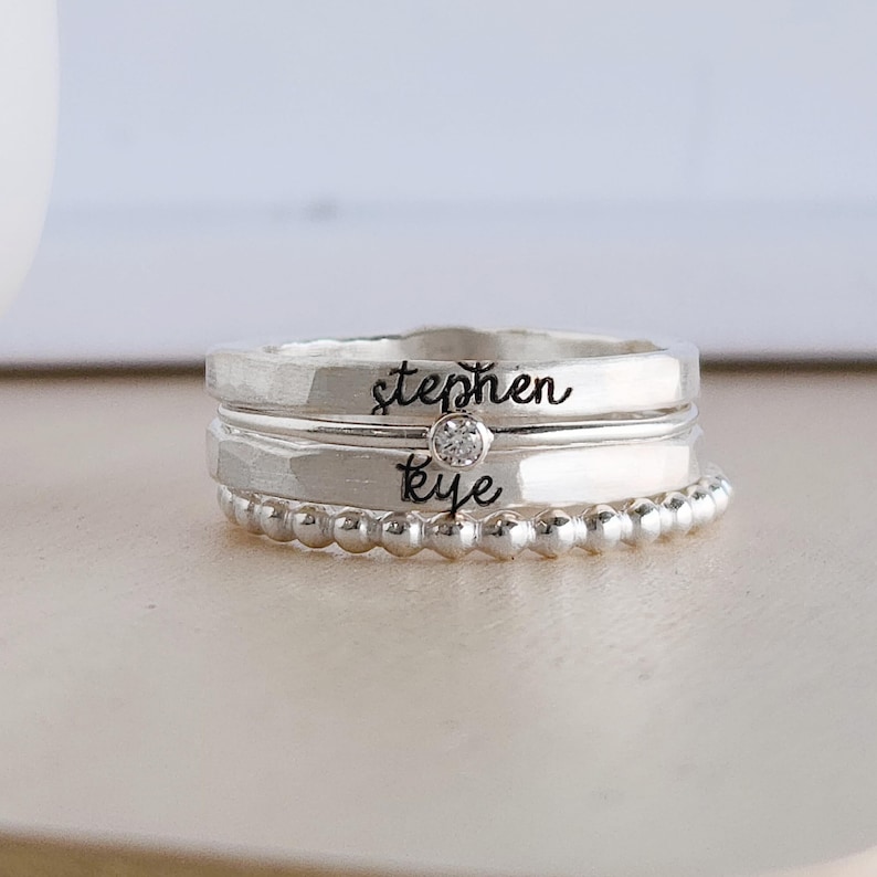 A stack of four silver-colored rings featuring engraved names stephen and kye, a beaded band, and a crystal solitaire ring.