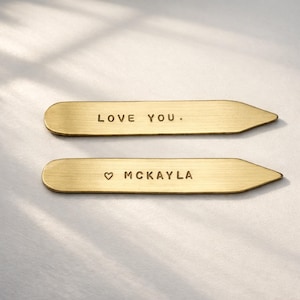 Two brass collar stays are side-by-side on a light surface, one engraved "LOVE YOU." and the other "♡ MCKAYLA."