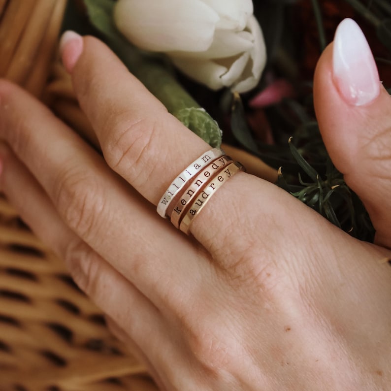 May include: Three stacked personalized rings with the names "William", "Kennedy", and "Audrey" engraved on them. The rings are worn on a person's finger.