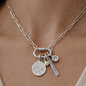 A silver paperclip chain necklace with a carabiner clasp holds four charms: a round disc with "OWEN" stamped on it, a small white pearl, a silver bar with "K" stamped on it, and a round clear gemstone in a bezel setting, worn by a person.