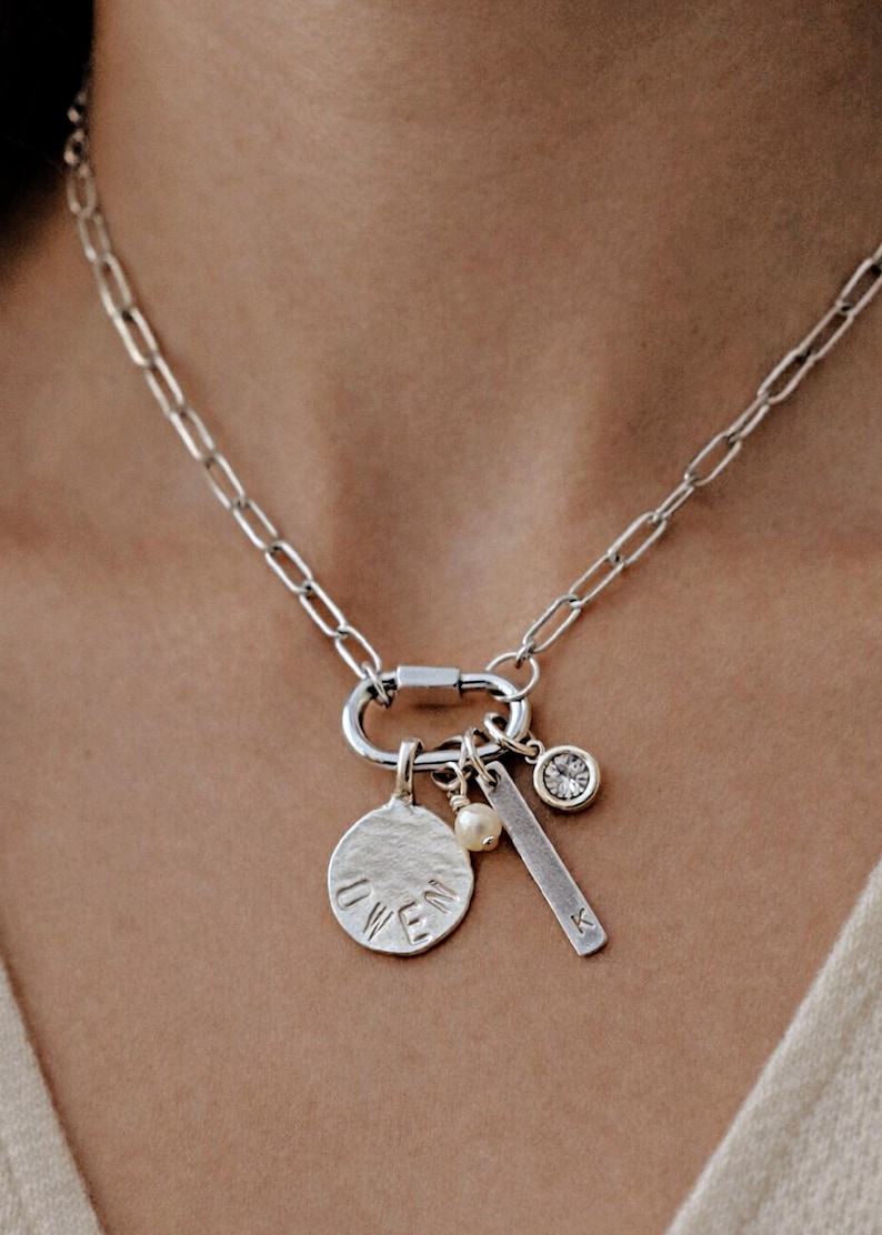 A silver paperclip chain necklace with a carabiner clasp holds four charms: a round disc with "OWEN" stamped on it, a small white pearl, a silver bar with "K" stamped on it, and a round clear gemstone in a bezel setting, worn by a person.