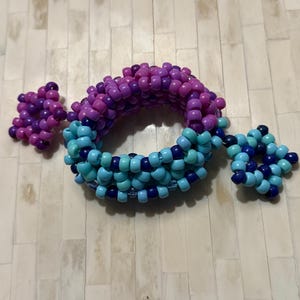 May include: A beaded bracelet in shades of purple, blue, and turquoise. The bracelet is circular with two star-shaped accents on either side. The beads are small and evenly spaced, creating a textured surface. The bracelet is likely handmade.