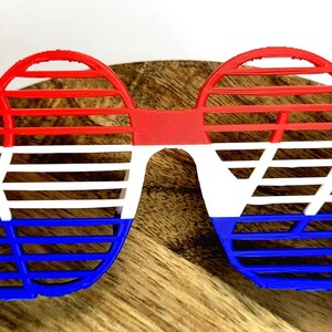May include: Red, white, and blue plastic sunglasses with a grid pattern. The sunglasses are shaped like a traditional pair of sunglasses.