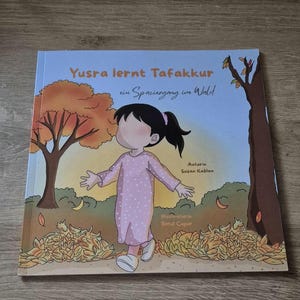 Könnte beinhalten: Ein Kinderbuch mit dem Titel Yusra lernt Tafakkur ein Spaziergang im Wald, das ein Kind mit Pferdeschwanz in einem rosa gepunkteten Kleid zeigt, das durch einen Wald mit orangefarbenen und braunen Herbstblättern am Boden geht.
