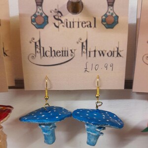 May include: A pair of blue and white mushroom earrings with gold hooks. The earrings are handmade and have a whimsical, fairy tale aesthetic. The price tag reads "Surreal Alchemy Artwork £10.99".