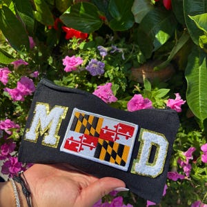 May include: Black rectangular pouch with the word "MOOD" in gold and white lettering. The pouch features a Maryland flag patch. The background includes pink and purple flowers and green foliage.