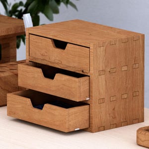 Könnte beinhalten: Ein kleiner Schreibtisch-Organizer aus hellbraunem Holz mit drei übereinanderliegenden Schubladen und Griffmulden. Die Einheit ist mit sichtbaren Steckverbindungen an den Seitenwänden konstruiert und bietet eine strukturierte Aufbewahrung.