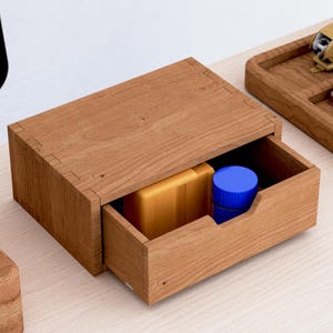 Könnte beinhalten: Ein rechteckiger Schreibtisch-Organizer aus Holz mit einer einzelnen Schublade. Im Inneren der offenen Schublade befinden sich ein gelbes rechteckiges Objekt und ein kleiner blauer zylindrischer Behälter. Das Holz hat eine natürliche Maserung.
