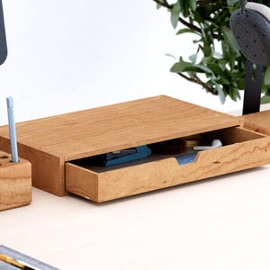 Könnte beinhalten: Ein rechteckiger Schreibtisch-Organizer aus Holz mit einer ausziehbaren Schublade und einer natürlichen Holzmaserung. Die Schublade ist teilweise geöffnet und zeigt kleine Büroartikel. Ein passender Stiftehalter aus Holz steht links.