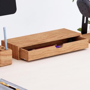 Könnte beinhalten: Ein rechteckiger Schreibtisch-Organizer aus Holz mit einer ausziehbaren Schublade und einem Griffausschnitt. Die hellbraune Holzmaserung ist auf der Oberfläche sichtbar. Die Schublade ist leicht geöffnet und zeigt einen roten und einen violetten Stift.